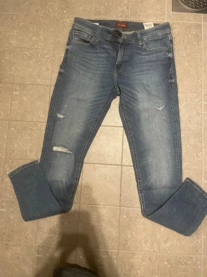 Blå slitna jeans från jack and jones - Säljer ett par blå jeans från Nudie Jeans med snygga slitningar på låret och knät. Klassisk femficksmodell med normal midja och raka ben. Jeansen har uppvikta benslut och är gjorda i mjuk denim med lite stretch för extra komfort.
