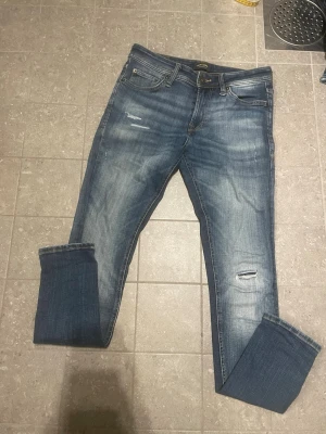 Blå slitna jeans från Jack & Jones - Säljer ett par blå jeans från Jack & Jones med snygga slitningar och tvättade detaljer. Klassisk femficksmodell med raka ben och uppvikta benslut. Jeansen har en skön passform och är tillverkade i bomull med lite stretch för extra komfort.