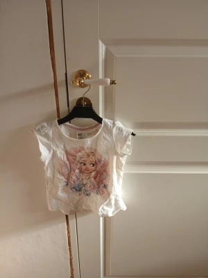 Vit topp med Elsa tryck H&M - Söt vit topp från H&M med korta ärmar och ett blommigt med elsa ifrån frost tryck.  Toppen har en klassisk rund halsringning och är gjord i mjuk bomull, perfekt för en avslappnad och trendig look.