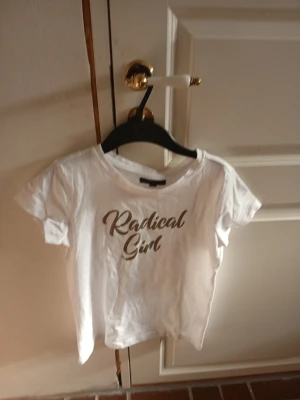 Vit t-shirt med texten Radical Girl - Vit t-shirt med korta ärmar och rund halsringning. På framsidan står det 'Radical Girl' i guldigt tryck. T-shirten har en enkel och clean design, perfekt för en avslappnad look.
