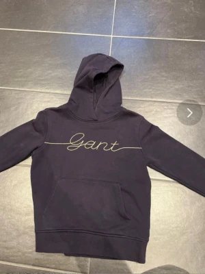 Mörkblå hoodie från Gant - Snygg mörkblå hoodie från Gant med guldfärgat broderat logotyp på bröstet. Klassisk känguruficka framtill och huva. Tillverkad i mjuk bomull för skön känsla. Perfekt för en avslappnad och trendig look.