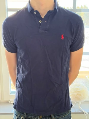 Marinblå pikétröja från Ralph Lauren - Klassisk marinblå pikétröja från Ralph Lauren med röd broderad logga på bröstet. Tröjan har korta ärmar, krage och knappar vid halsen. Tillverkad i bomull för en bekväm och avslappnad stil.