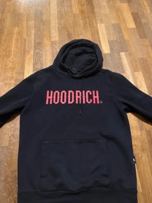 Svart HOODRICH hoodie med röd logga - Svart hoodie från HOODRICH med stor röd logga på bröstet. Klassisk passform med huva och dragsnöre samt en stor magficka framtill. Tillverkad i mjukt material som känns skönt mot huden. Perfekt för en streetwear-look.