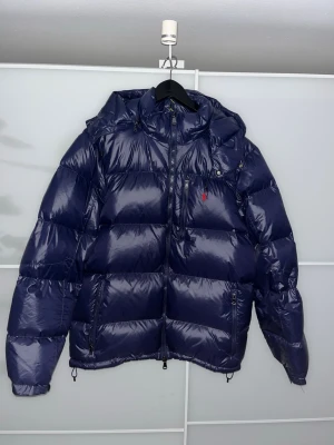 Mörkblå pufferjacka från Polo Ralph Lauren - Snygg mörkblå pufferjacka från Polo Ralph Lauren med blank finish och klassisk röd logga på bröstet. Jackan har huva, dragkedja framtill och flera fickor. Perfekt för kalla vinterdagar och med en extra lång vinter i år.🛑 Tveka inte med att höra av dig vid fundering såsom pris eller fler bilder.