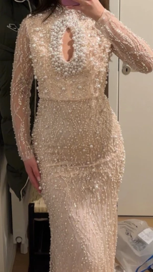 Lång ljus pärlprydd klänning HELT NY - Elegant långklänning i ljus beige ton, täckt med pärlor och paljetter. Klänningen har långärmade meshärmar, hög halsringning och en dekorativ öppning framtill. Figurnära passform och exklusiv look, perfekt för speciella tillfällen.