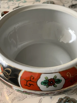 Färgglad asiatisk porslinsskål med lock - Säljer en rund porslinsskål med lock, dekorerad i rött, svart, vitt och guld. Skålen har blommönster och asiatiska motiv runt om samt gulddetaljer på locket. Perfekt som dekoration eller serveringsskål. Materialet är glansigt porslin.