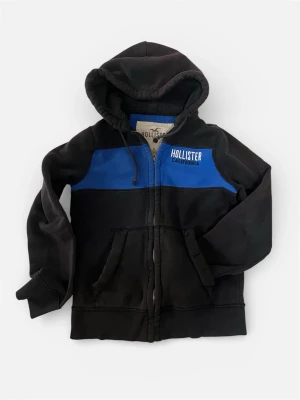 Svart och blå hoodie från Hollister - Snygg svart hoodie med blå panel och dragkedja från Hollister. Hoodien har huva med dragsko, fickor framtill och Hollister California-tryck på bröstet. Perfekt för en avslappnad och cool stil. Tillverkad i mjukt bomullsmaterial.