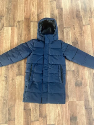 Mörkblå dunjacka från Only & Sons - Snygg mörkblå dunjacka från Only & Sons med huva och raka, quiltade paneler. Jackan har två svarta dragkedjefickor framtill och en hög krage som skyddar mot kyla. Perfekt för kalla vinterdagar och har en stilren, lång modell. Nytt skick