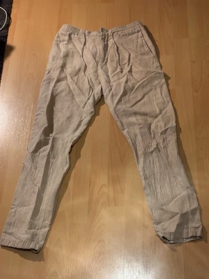 Beiga linnebyxor från J.Lindeberg - Snygga och avslappnade beiga linnebyxor från J.Lindeberg. Byxorna har resår i midjan med dragsko och raka ben. Tillverkade i ett lätt och luftigt linnematerial som passar perfekt för en chill och stilren look.