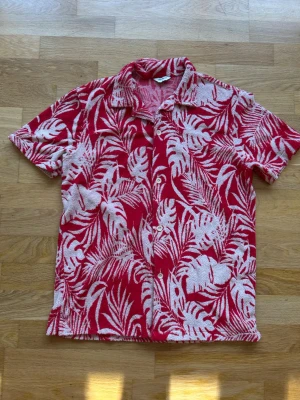 Röd blommig skjorta med knappar från East west - Snygg röd skjorta med vita blommönster och blad, klassisk krage och knäppning framtill. Kortärmad och perfekt för sommarens varma dagar. Skjortan har en avslappnad passform och är tillverkad i ett mjukt material.
