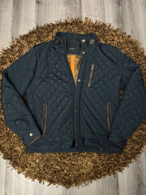 Mörkblå quiltad jacka från Saki - Quiltad mörkblå jacka från Saki med bruna detaljer vid dragkedjor och krage. Jackan har hög krage med tryckknapp, tre fickor med dragkedja och snyggt rutmönster. Perfekt för kyligare dagar och ger en stilren look.