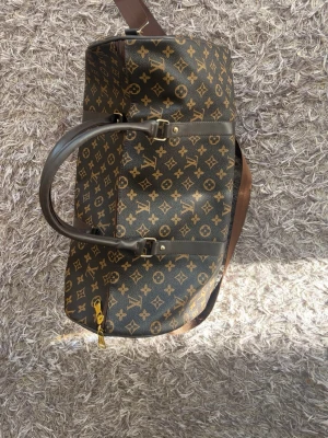 Louis Vuitton monogram weekendbag - Säljer en klassisk brun weekendbag från Louis Vuitton med det ikoniska monogrammönstret i guld. Väskan har två robusta handtag, justerbar axelrem och guldfärgade metalldetaljer. Perfekt för resor när du vill resa med stil.