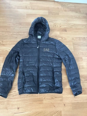 Svart dunjacka från EA7 Emporio Armani - Snygg svart dunjacka från EA7 Emporio Armani med huva och dragkedja framtill. Jackan har diskret logga på bröstet och två sidofickor. Lättviktig och quiltad design som ger en modern look. Perfekt för kalla vinterdagar. Nypris ca 1200kr mitt pris 399kr.