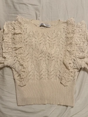 Vit stickad topp med volang från Zara - Supersöt vit stickad topp från Zara med korta puffärmar och dekorativa virkade volanger längs axlarna. Tröjan har ett fint hålmönster och ribbad nederkant. Perfekt för dig som gillar romantisk och trendig stil.