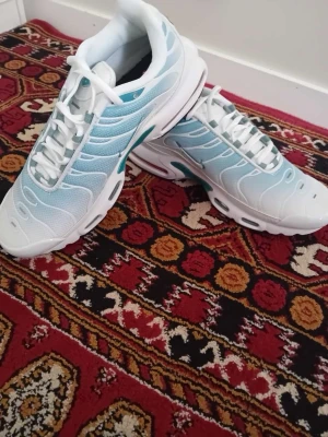 Nike Air Max Plus vita/blå sneakers - Nike Air Max Plus sneakers i vitt och ljusblått med klassisk vågig design och synlig Air-dämpning i sulan. Ovandelen är i mesh och syntet, med vita snören och detaljer i turkos. Perfekta för dig som gillar sportig och fräsch stil.
