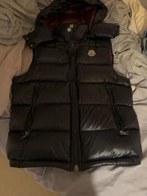 Äkta mörk blå Moncler väst  - Snygg mörkblå Moncler väst från Moncler med avtagbar huva och två dragkedjefickor framtill. Västen har en glansig finish och klassisk Moncler-logga på bröstet. Perfekt för lager-på-lager och streetstyle.