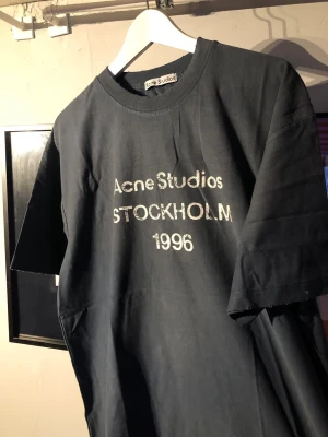 Svart Acne Studios t-shirt med tryck - Cool svart t-shirt från Acne Studios med vit text 'STOCKHOLM 1996' på bröstet. Klassisk rund hals och korta ärmar. Perfekt för dig som gillar stilrena och trendiga plagg med streetkänsla.