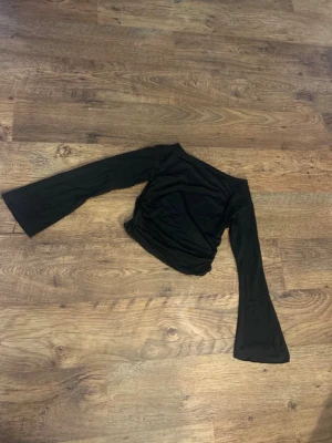 Svart långärmad topp med utsvängda ärmar - En svart långärmad topp med utsvängda ärmar och scrunsh vid midjan med en enkel, stilren design. Toppen har en tight passform och är tillverkad i ett tunt syntetmaterial som ger en lätt och smidig känsla. Perfekt för att skapa en cool och trendig look. Storlek L passar även M
