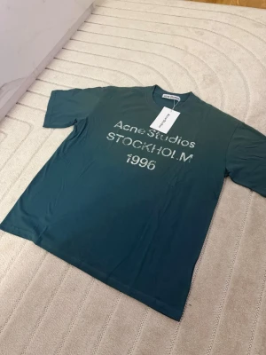 Blå Acne Studios t-shirt S - Snygg blå t-shirt från Acne Studios med vit text 'Acne Studios STOCKHOLM 1996' på bröstet. Klassisk rund hals och korta ärmar. Tillverkad i mjuk bomull för en skön känsla och avslappnad passform. Perfekt för dig som gillar stilrena och trendiga plagg.
