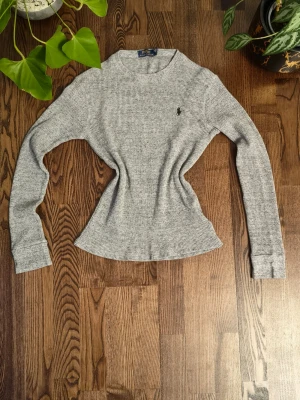 Grå waffleknitt långärmad topp Polo Ralph Lauren - Snygg grå våfflad långärmad topp från Polo Ralph Lauren med rund halsringning och liten broderad logga på bröstet. Figurnära passform och mjukt material som känns skönt mot huden. Perfekt att styla med jeans eller kjol.