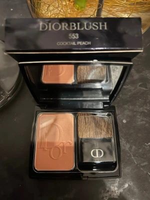 Diorblush 553 Cocktail Peach rouge - Diorblush i nyansen 553 Cocktail Peach, en persikofärgad rouge med lätt skimrig finish. Kommer i en lyxig svart dosa med spegel och tillhörande borste med Dior-logga. Perfekt för att ge kinderna en fräsch och naturlig glow.