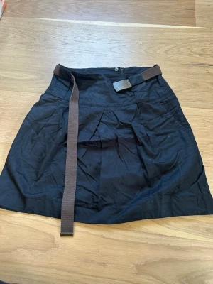 Svart kjol med bälte från Zara - Snygg svart kortkjol från Zara med brunt tygbälte och dekorativt spänne. Kjolen har dragkedja i sidan och lätt utställd passform. Perfekt att styla med en enkel topp eller hoodie för en chill look.