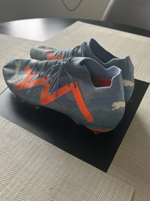 Blå-orange Puma fotbollsskor - Säljer ett par snygga blå fotbollsskor från Puma med orangea detaljer och vita Puma-loggor. Skorna har snörning och en strumpliknande krage för extra komfort. Perfekta för dig som vill sticka ut på planen med både stil och funktion. Använda endast 1 säsong