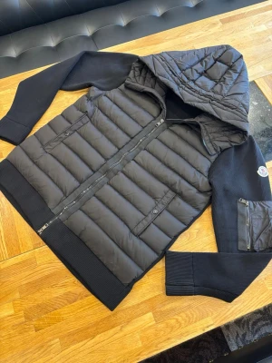 Moncler Cardigan  - Snygg svart dunjacka från Moncler med vadderad kropp och stickade ärmar. Jackan har en stor huva, två framfickor med dragkedja och en Moncler-logga på ärmen. Perfekt för kalla vinterdagar och riktigt stilren look.