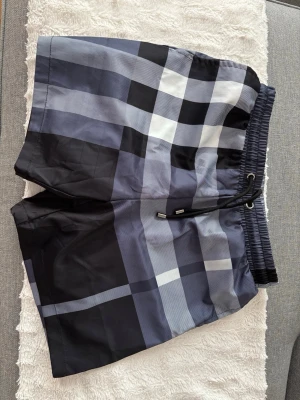 Rutiga badbyxor från Burberry - Snygga badbyxor från Burberry i klassiskt rutigt mönster i svart, vitt och grått. De har elastisk midja med dragsko och innerbyxa i mesh för extra komfort. Perfekta för stranden eller poolen, tillverkade i snabbtorkande material.