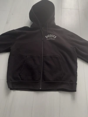 Svart zip hoodie från Hoodrich - Säljer en svart hoodie med dragkedja från Hoodrich. Stor vit logga och text 'Welcome to the family' på både bröstet och ryggen. Hoodien har huva med dragsko och stora fickor framtill. Perfekt för en streetwear-look.