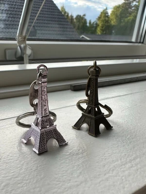 Eiffeltornet nyckelringar Paris - Två nyckelringar formade som Eiffeltornet, en i silverfärgad metall med texten 'FRANCE' och en i mörk bronsfärgad metall med texten 'PARIS'. Perfekta accessoarer för dig som gillar Paris eller vill ha en unik detalj på nyckelknippan.