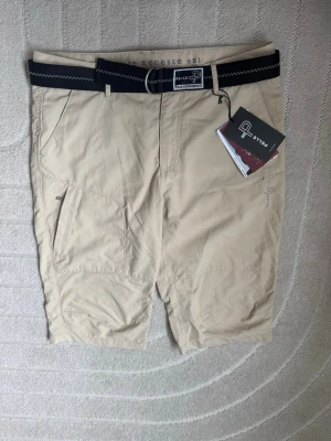 Beige shorts från Pelle P med bälte - Snygga beige shorts från Pelle P med svart bälte. Tillverkade i snabbtorkande material, perfekta för aktiva dagar. Klassisk passform och diskret logga på benet. Passar dig som gillar funktion och stil.