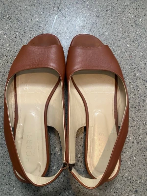 Bruna lädersandaler från Hobbs - Snygga bruna sandaler från Hobbs i äkta läder med öppen tå och slingback-rem. Skorna har en stilren design med beige innersula och låg klack, perfekta för en trendig och avslappnad look.