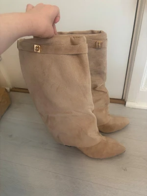 Beige slouchy stövlar i mocka-look - Säljer ett par beige slouchy stövlar med spetsig tå och låg klack. Stövlarna har en mjuk mocka-liknande yta och dekorativa guldfärgade knappar upptill. Perfekta för dig som vill ha en trendig och avslappnad stil.