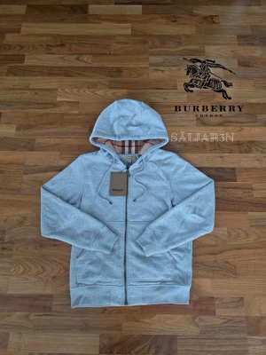 Grå Burberry Hoodie - Letar du efter en stilren och lyxig hoodie? Denna helt nya grå Burberry-hoodie kombinerar elegans med hög komfort. Den har Burberrys ikoniska rutmönster i fodret och en diskret logga på bröstet för en exklusiv touch. Tillverkad av mjukt, slitstarkt material och utrustad med dragkedja samt huva – både praktisk och stilfull.  📌 Skick: Ny med taggar. 📦 Snabb leverans – skickas samma dag (om ej hinder)