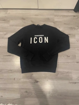 Svart sweatshirt från Dsquared2 ICON - Säljer en svart sweatshirt från Dsquared2 med stor vit 'ICON'-logga på bröstet. Tröjan har rund halsringning, långa ärmar och breda muddar vid ärmslut och nederkant. Perfekt för dig som gillar streetwear och vill sticka ut.