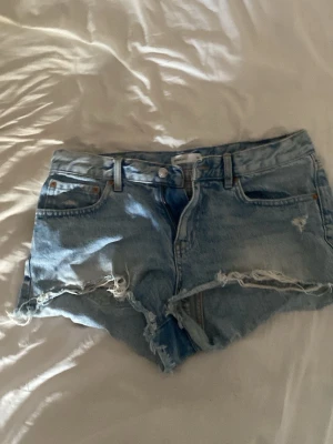 Ljusblåa denimshorts med slitningar - Säljer ett par ljusblåa denimshorts med råa kanter och slitna detaljer. Klassisk femficksmodell med knapp och dragkedja framtill. Perfekta för varma dagar och en avslappnad stil.