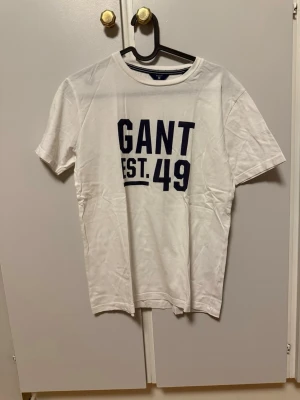 GANT t-shirt - Vit t-shirt från GANT med mörkblå text 'GANT EST. 49' på bröstet. Klassisk rund halsringning och korta ärmar. Tillverkad i mjuk bomull för en bekväm känsla och enkel stil. Storlek 170 cm