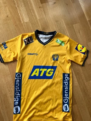 Gul svensk matchtröja från Craft - Säljer en gul matchtröja från Craft med flera sponsorer som ATG, Gjensidige och XL Bygg. Tröjan har blå och vita detaljer, V-ringad krage och tryckta loggor. Perfekt för dig som gillar svensk handboll eller vill ha en snygg supportertröja.