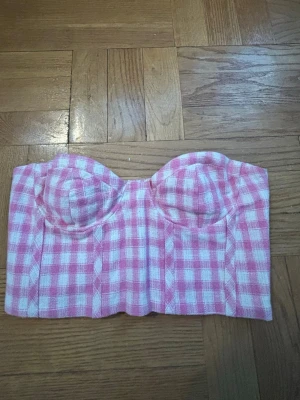 Rutig rosa korsett topp - Supersöt korsett topp i rosa och vitt med rutigt mönster. Toppen är axelbandslös och har vadderade kupor samt smockad rygg för extra komfort. Perfekt för sommarens alla tillfällen! 