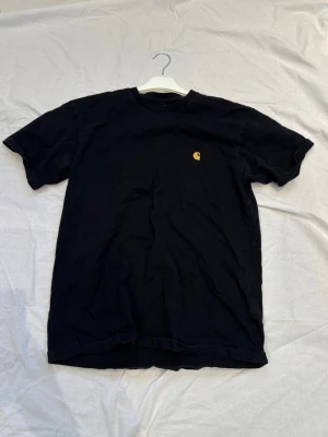 Svart Carhartt t-shirt med logga - Svart t-shirt från Carhartt med klassisk gul logga på bröstet. T-shirten har rund halsringning och korta ärmar. Perfekt för en avslappnad och stilren look.
