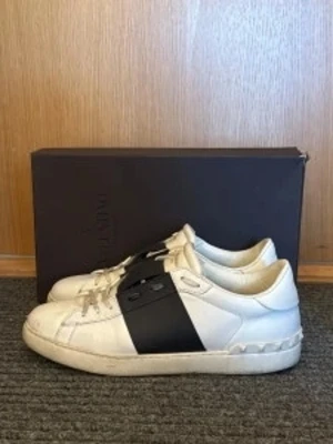Valentino vita sneakers med svart band - Snygga vita sneakers från Valentino med ett brett svart band över snörningen. Skorna har klassisk låg profil och är tillverkade i läder med detaljer i vitt och svart. Perfekta för dig som vill ha en stilren och trendig look.