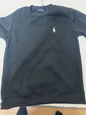 Svart sweatshirt från Polo Ralph Lauren - Klassisk svart sweatshirt från Polo Ralph Lauren med rund halsringning och den ikoniska vita logobrodyrn på bröstet. Tröjan har ribbade muddar vid ärmslut och nederkant. Perfekt för en clean och stilren look.