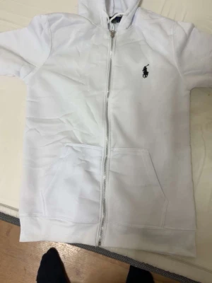 Vit zip hoodie från Ralph Lauren - Snygg vit hoodie med dragkedja från Ralph Lauren. Klassisk design med huva, fickor framtill och den ikoniska svarta loggan broderad på bröstet. Tillverkad i mjukt bomullsmaterial som känns skönt mot huden. Perfekt för en clean och stilren look.