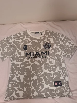 BAPE x Inter Miami camo t-shirt - Säljer en limited edition BAPE x Inter Miami t-shirt med grått och vitt camomönster. Stor MIAMI-logga och BAPE-ansikte på bröstet, samt stort BAPE 93-tryck på ryggen. Skön passform och cool streetwear-vibe, perfekt för dig som gillar unika samarbeten.