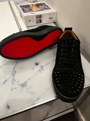 Svarta Louboutin sneakers med nitar - Säljer ett par svarta sneakers från Christian Louboutin med klassisk röd sula och svarta nitar på tån. Skorna har snörning, låg profil och är tillverkade i mocka och läder. Perfekta för dig som vill sticka ut med en edgy och lyxig stil.