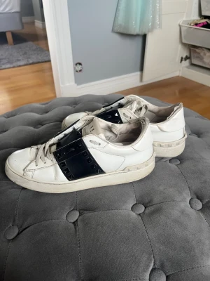 Vita sneakers från Valentino - Snygga vita sneakers från Valentino med svart band och nitar på sidan. Skorna har klassisk låg profil, vita snören och en chunky sula. Tillverkade i läder med detaljer i svart och vitt. Perfekta för dig som gillar exklusiv streetstyle.
