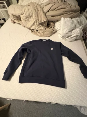 Mörkblå sweatshirt från Moncler - Säljer en stilren mörkblå sweatshirt från Moncler med rund halsringning och diskret Moncler-logga på bröstet. Tröjan har långa ärmar och ribbade muddar vid ärmslut och nederkant. Perfekt för en clean och avslappnad look.