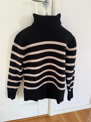 Svart- och beige randig polotröja - Stickad polotröja i svart med breda beige ränder, från Bondelid. Tröjan har hög, ribbad polokrage och ribbade muddar vid ärmslut och nederkant. Perfekt för kalla dagar och enkel att matcha med jeans eller kjol.