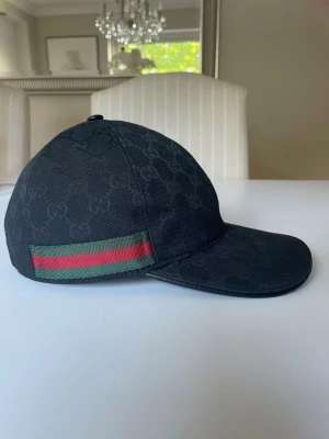 Svart keps från Gucci med GG-mönster - Snygg svart keps från Gucci med klassiskt GG-monogram över hela kepsen. På sidan finns en ikonisk grön och röd rand. Kepsen har böjd skärm och är tillverkad i canvasmaterial. Perfekt för dig som vill ha en stilren och lyxig accessoar.